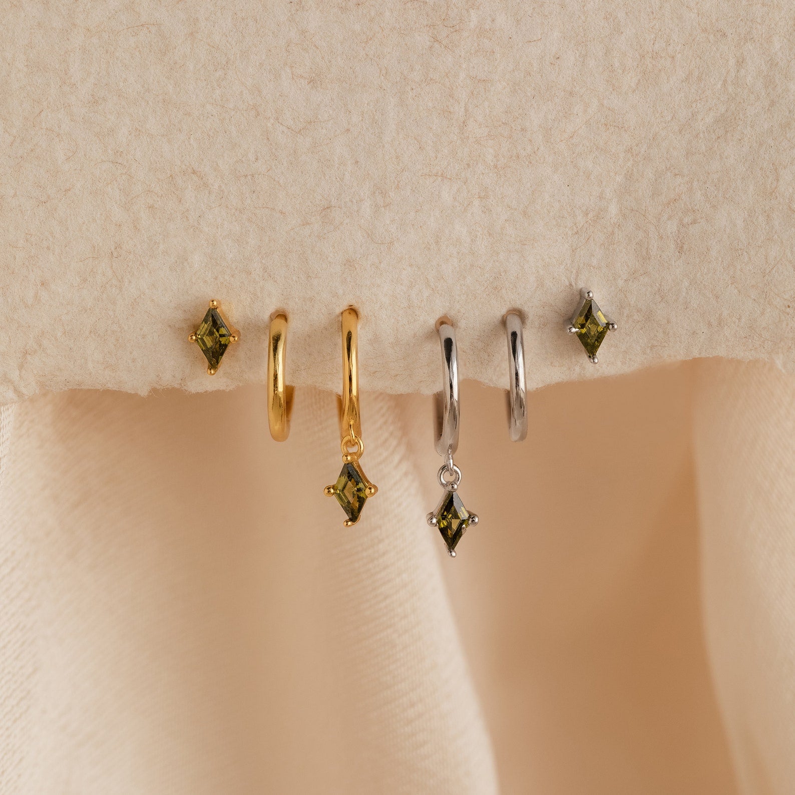 The Peridot Earrings Set in 18K Gold features two pairs of gold and silver hoop stacking earrings with green square gemstones, displayed on a beige textured surface.