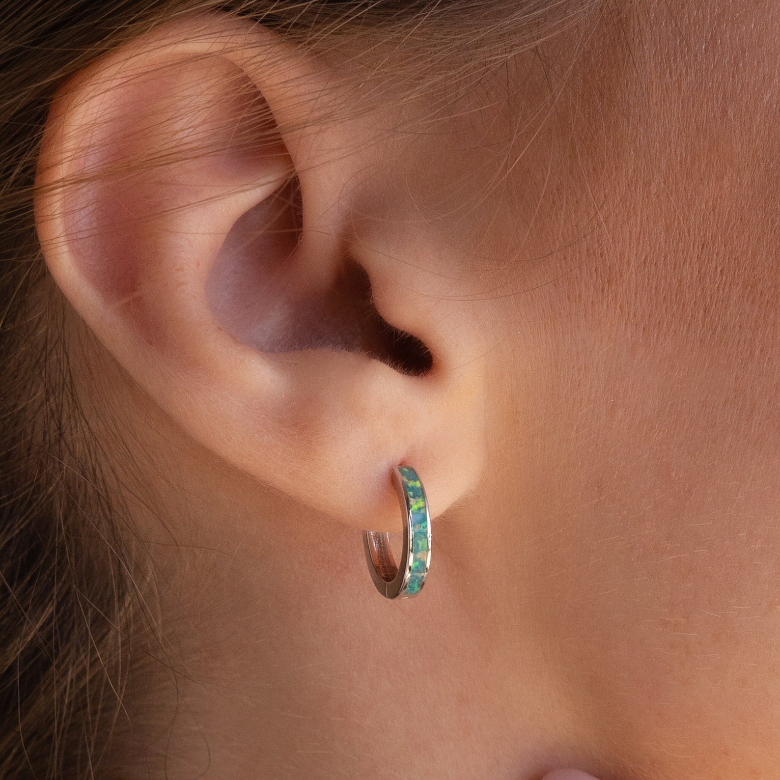 Close-up of a person’s ear adorned with small hoop earrings featuring a blue-green inlay, similar to the elegant Green Opal Inlay Hoops.