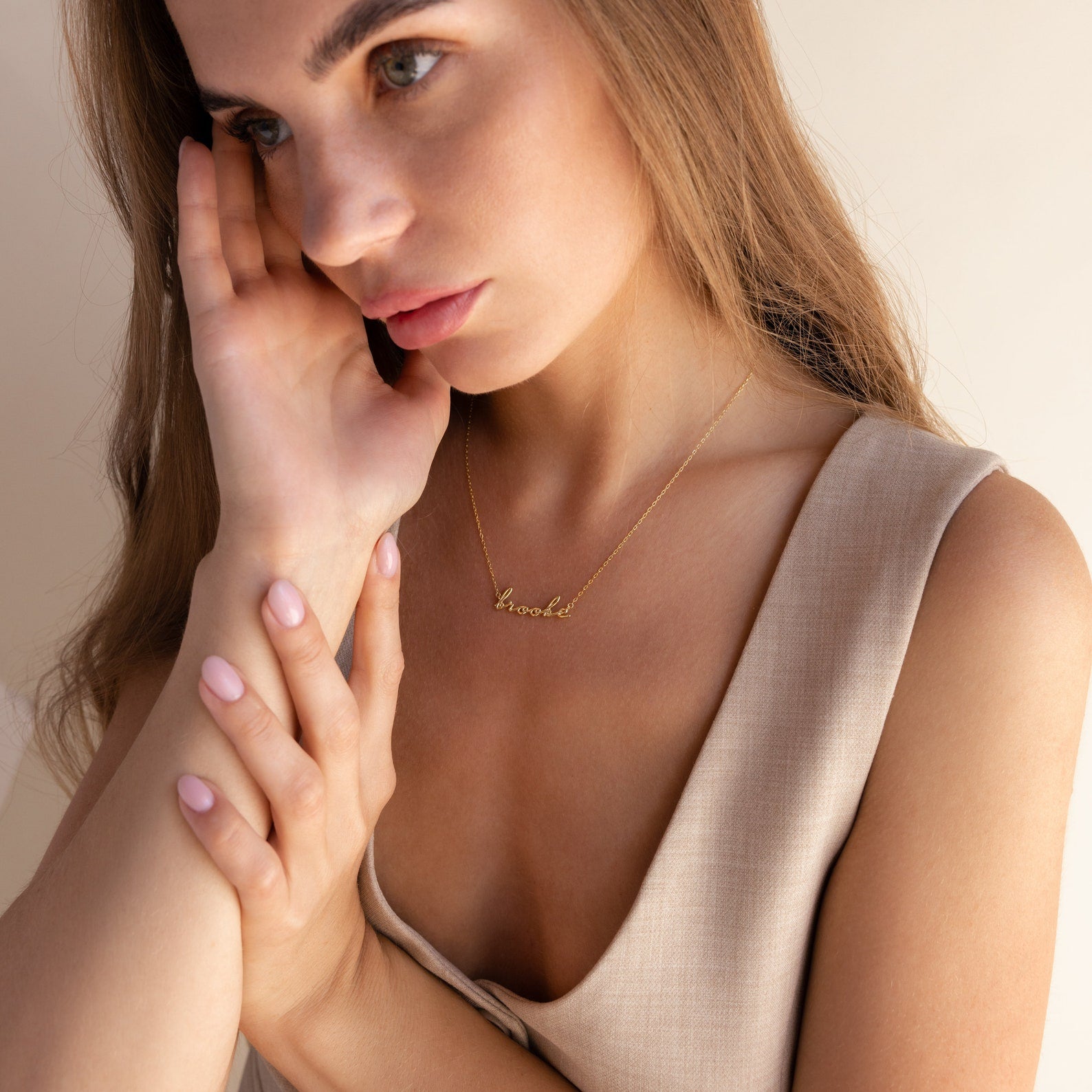 Woman with long hair wears a sleeveless beige top and gently touches her face while showcasing the Dainty Bubble Name Necklace in gold.