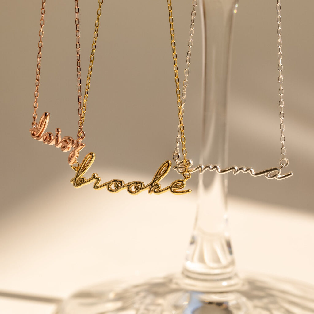 Three Dainty Bubble Name Necklaces in gold, rose gold, and silver hang on a glass stand in sunlight—a beautiful showcase of personalized nameplate jewelry.