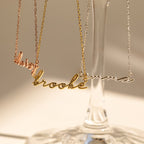 Three Dainty Bubble Name Necklaces in gold, rose gold, and silver hang on a glass stand in sunlight—a beautiful showcase of personalized nameplate jewelry.