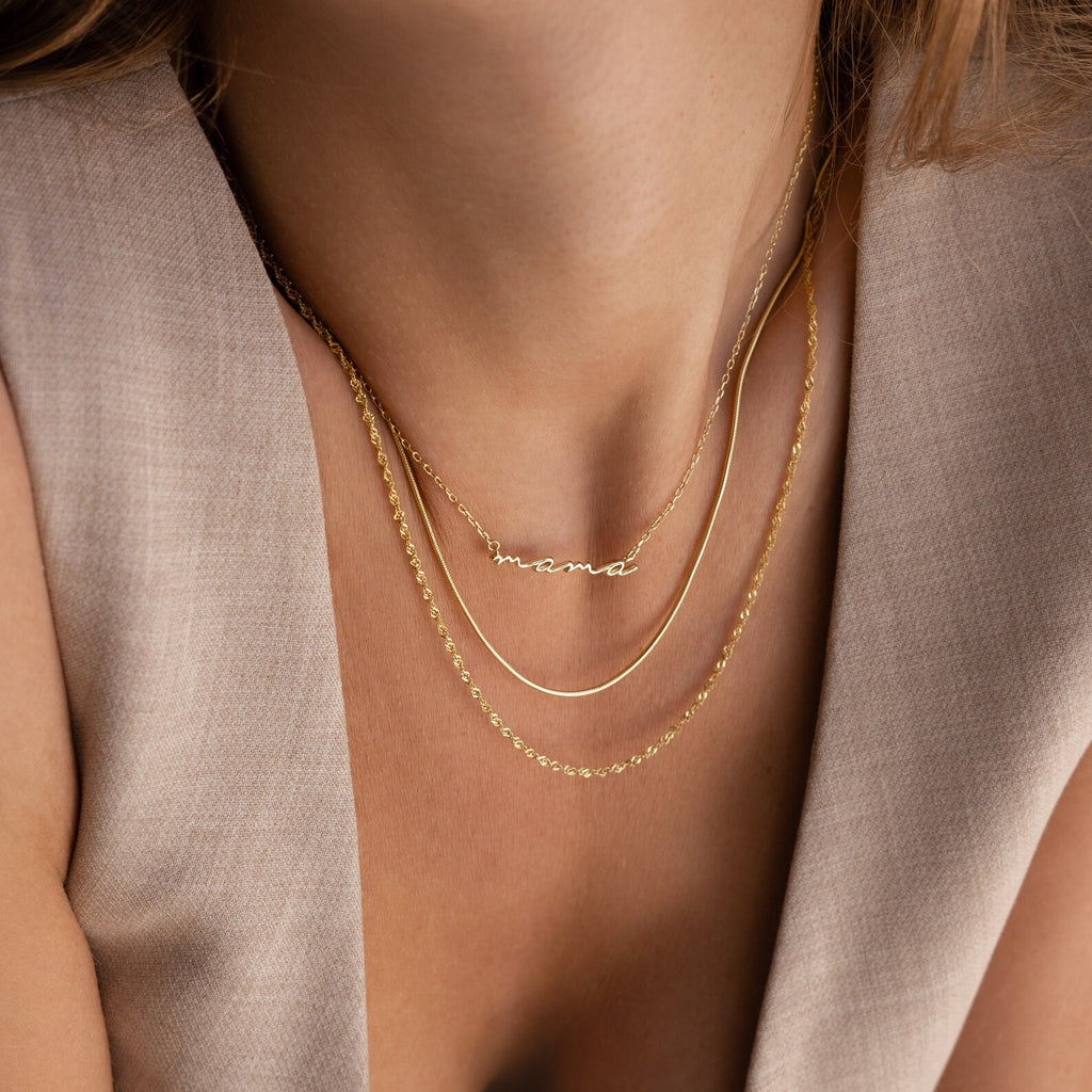 Close-up of a woman wearing three gold necklaces, including the Dainty Bubble Name Necklace with a "mama" pendant, styled with a beige sleeveless top.