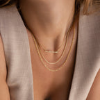 Close-up of a woman wearing three gold necklaces, including the Dainty Bubble Name Necklace with a "mama" pendant, styled with a beige sleeveless top.