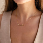 A woman wears the Dainty Bubble Name Necklace personalized with "Brooke" in gold cursive, styled with a beige sleeveless top.