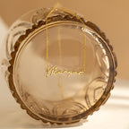 The Tiny Venice Name Necklace draped over the rim of a transparent, scalloped glass jar—this personalized piece adds a delicate nameplate charm to any setting.