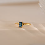 The Blue Topaz Emerald Ring, a vintage gold accessory with a rectangular blue topaz, is elegantly showcased on a soft beige surface.