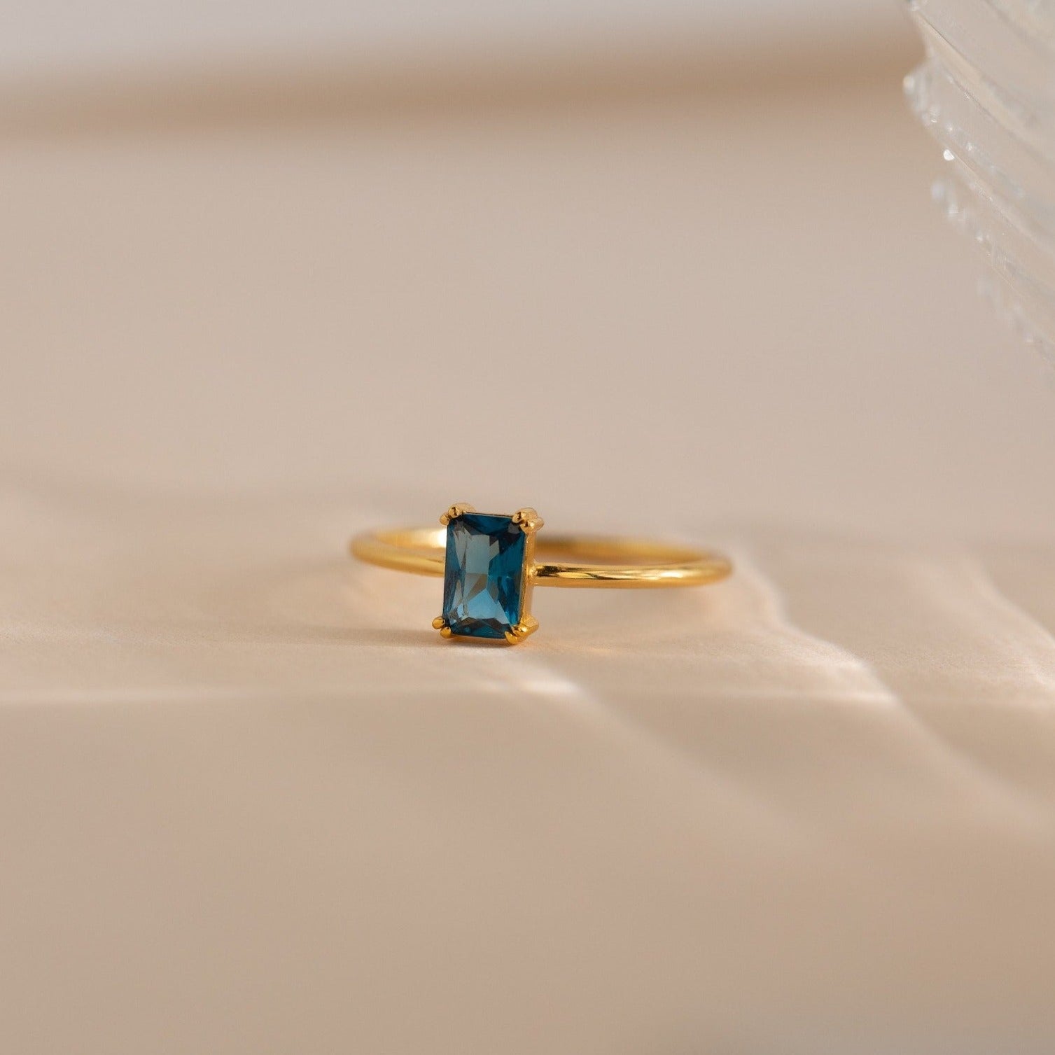 The Blue Topaz Emerald Ring, a vintage gold accessory with a rectangular blue topaz, is elegantly showcased on a soft beige surface.