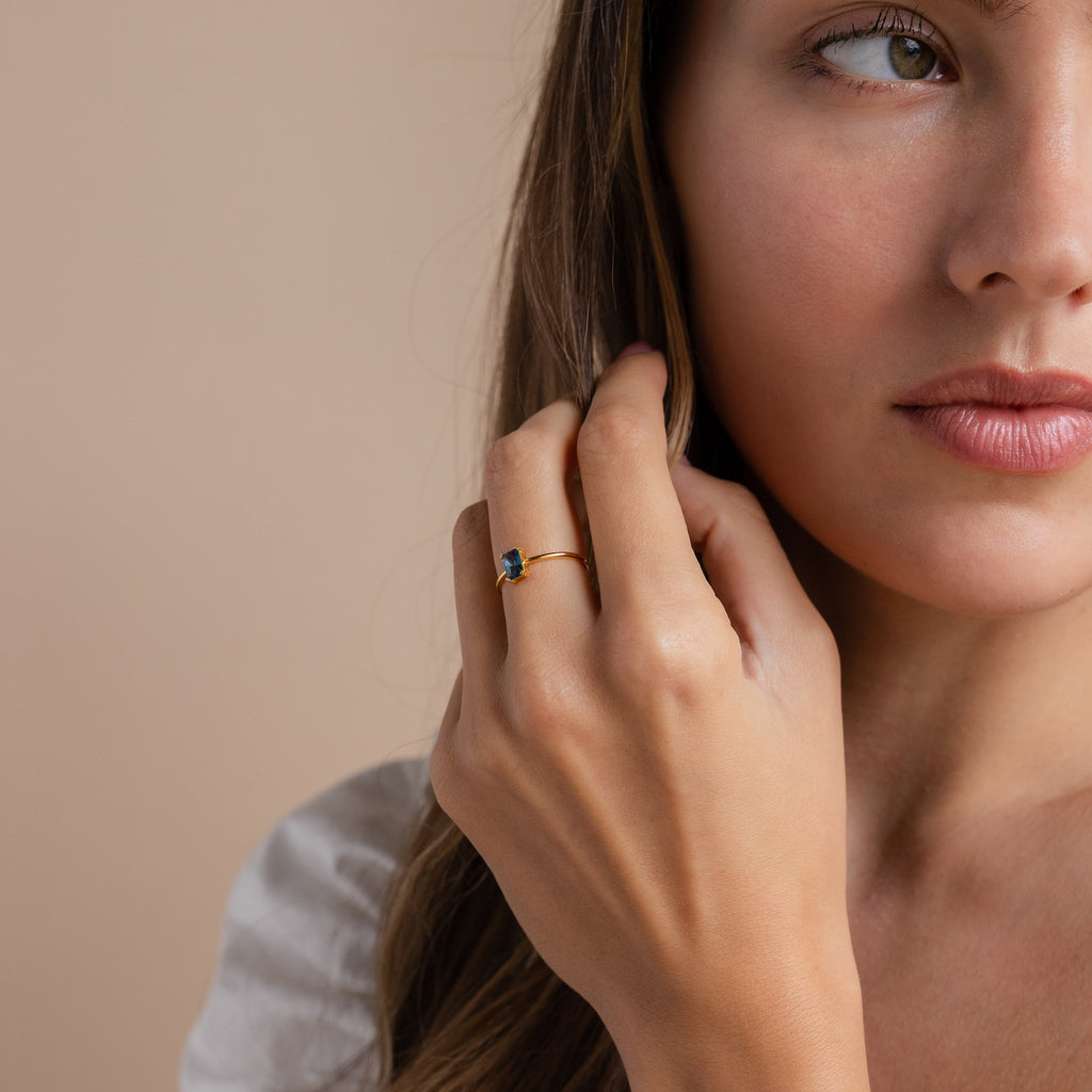 A woman with long brown hair gently touches her face, wearing a vintage-inspired Blue Topaz Emerald Ring featuring a striking blue gemstone.