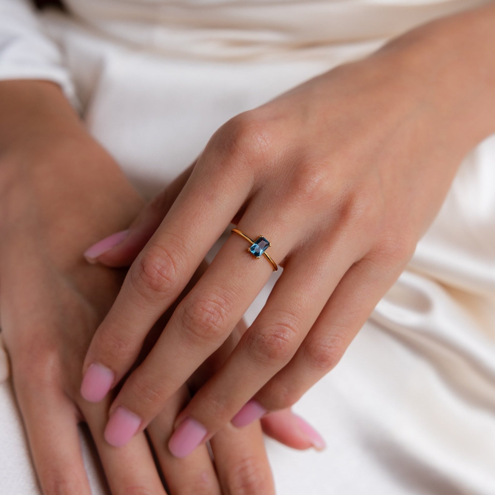 A hand with pink nails wearing a vintage accessory—a gold Blue Topaz Emerald Ring—rests elegantly on white fabric.