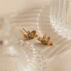 Aquamarine Marquise Studs with gold and blue floral design rest on cream fabric beside a clear glass object—ideal as a bridesmaids gift or for adding elegance to any special occasion.