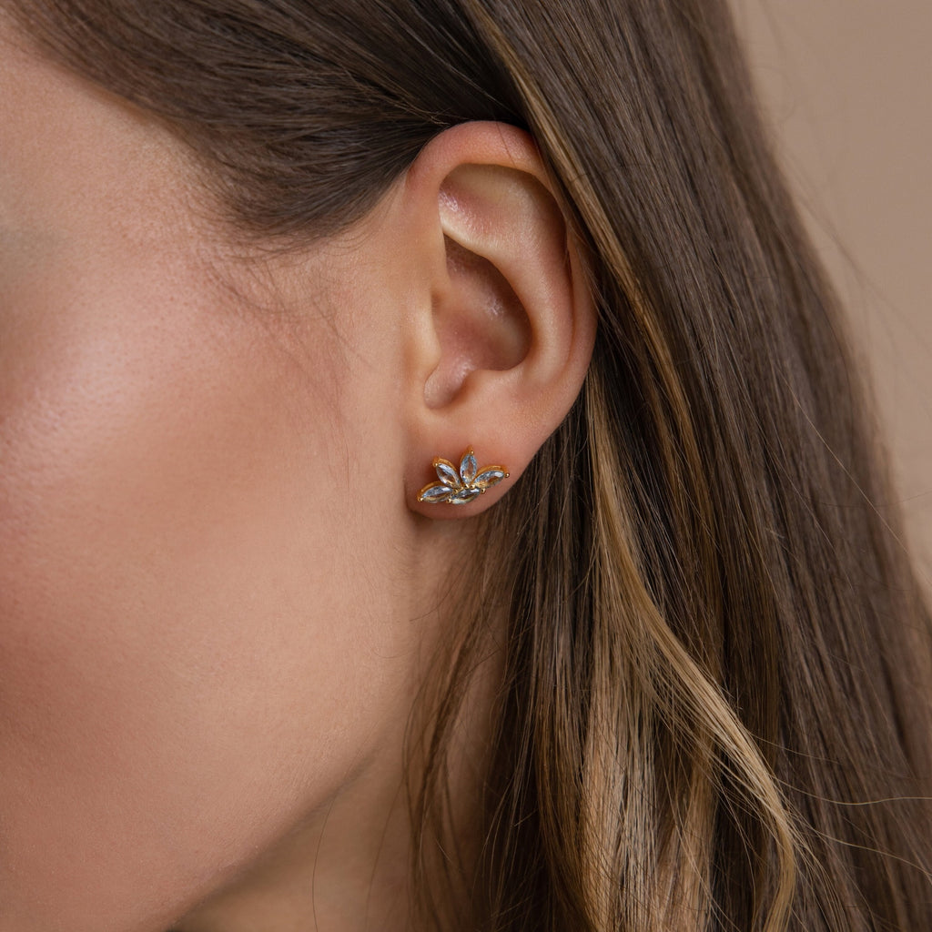 A woman's ear adorned with elegant gold floral earrings featuring small gemstones—an ideal bridesmaid gift or a complement to the Aquamarine Marquise Studs.