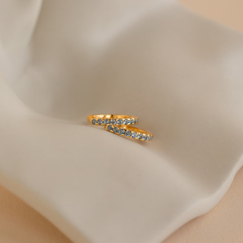 Two gold rings with small blue gemstones rest on cream-colored fabric—perfect for pairing with your Aquamarine Pave Huggies for an elegant ear stack.