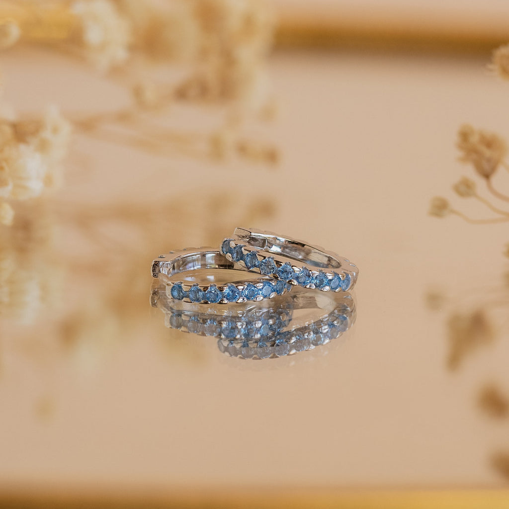 Two silver rings with blue gemstones rest on a reflective surface, accented by soft florals in the background, showcasing the refined elegance of Aquamarine Pave Huggies.