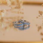 Two silver rings with blue gemstones rest on a reflective surface, accented by soft florals in the background, showcasing the refined elegance of Aquamarine Pave Huggies.