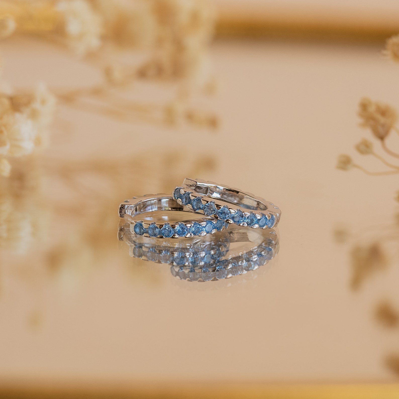 Two silver rings with blue gemstones rest on a reflective surface, accented by soft florals in the background, showcasing the refined elegance of Aquamarine Pave Huggies.