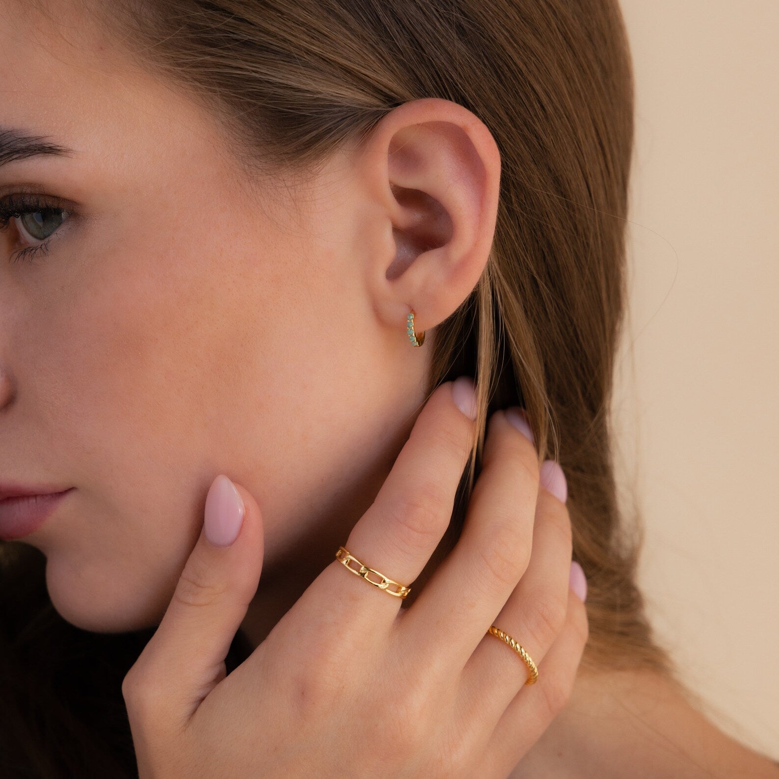 Close-up of a woman's ear wearing Garden Green Opal Huggies in an elegant earring stack with gold rings and manicured nails touching her face.