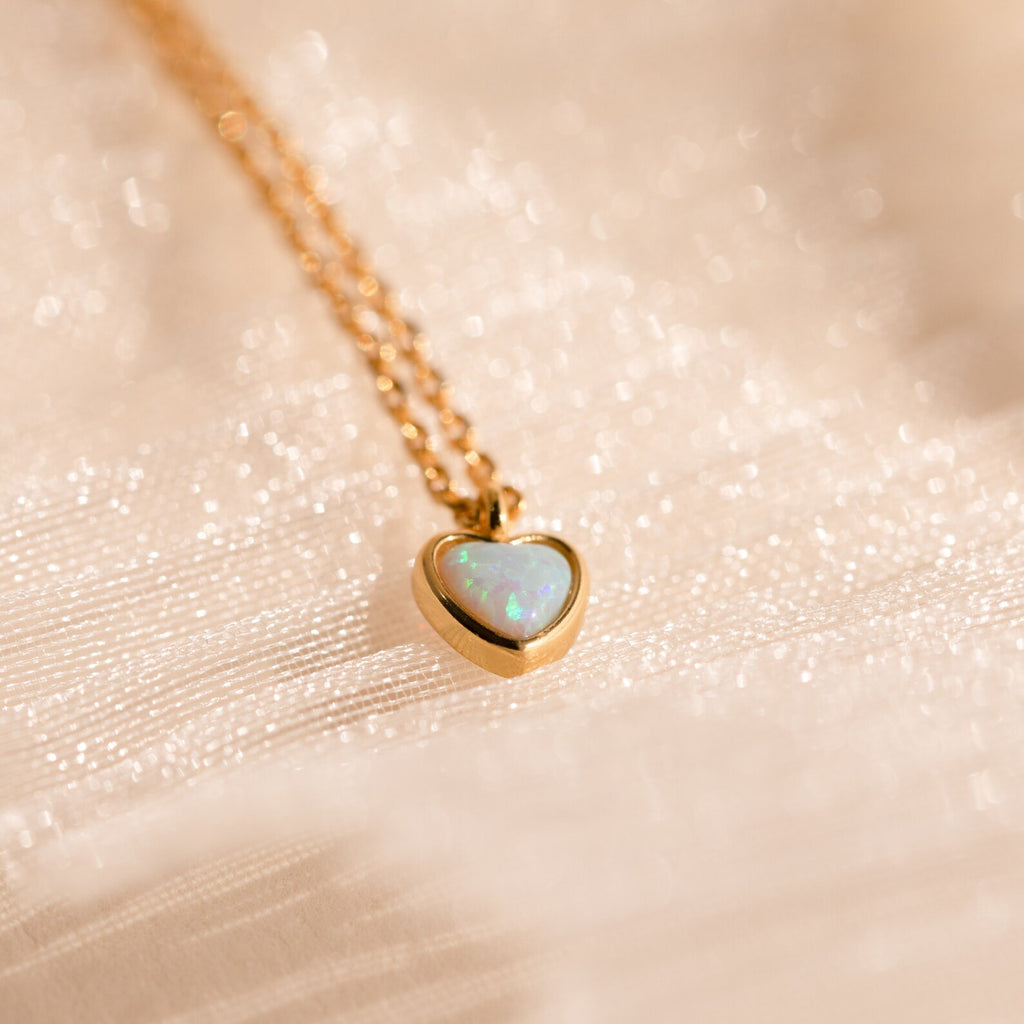 Tiny Opal Heart Necklace: A delicate gold necklace with a heart-shaped opal pendant, presented on soft, shimmery fabric for an elegant accent.