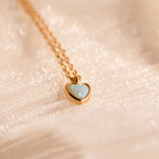 Tiny Opal Heart Necklace: A delicate gold necklace with a heart-shaped opal pendant, presented on soft, shimmery fabric for an elegant accent.