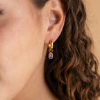 Close-up of a gold finish Dangling Gemstone Hoop earring with a rectangular purple stone, worn on a woman with curly hair. Features removable emerald-cut gemstones.
