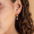 Close-up of a woman's ear wearing Dangling Emerald Birthstone Hoops—a gold hoop earring with a rectangular purple gemstone pendant.
