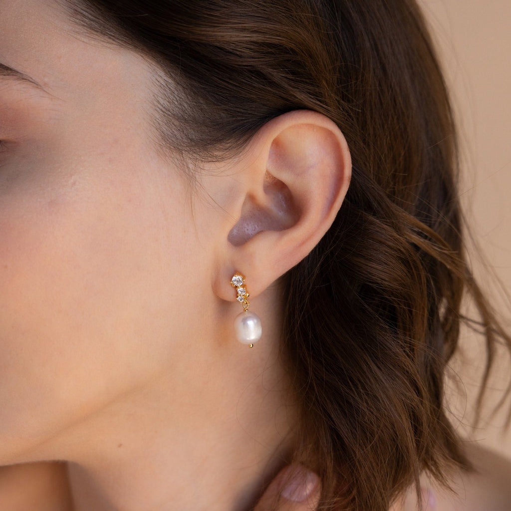 Close-up of a model wearing one Millie Drop Earring in gold, featuring a dangling pearl and sparkling stones.