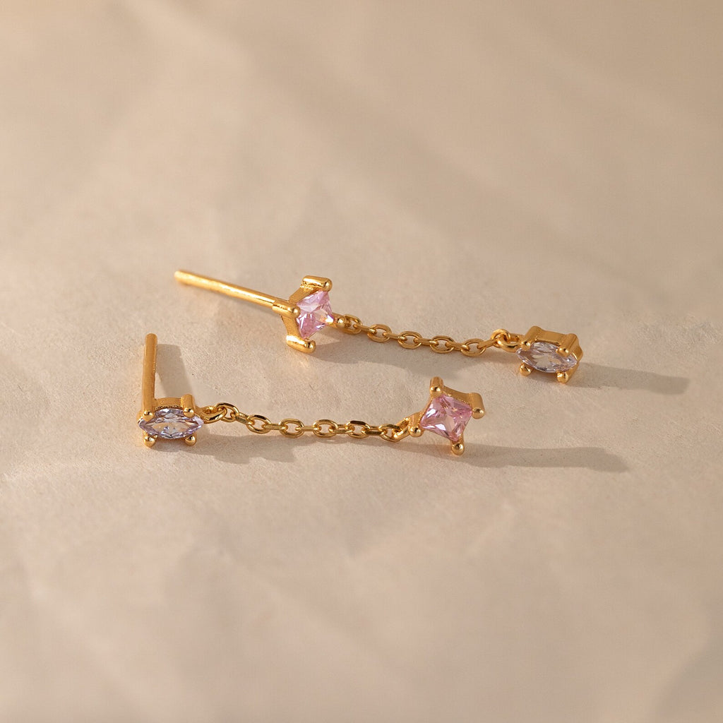 The Mixed Crystal Drop Earrings feature gold chain drops with pink and lavender gemstones, creating a minimalist style perfect for any occasion. These elegant dangling studs are shown on a light beige surface.