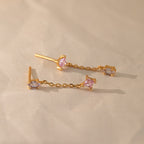 The Mixed Crystal Drop Earrings feature gold chain drops with pink and lavender gemstones, creating a minimalist style perfect for any occasion. These elegant dangling studs are shown on a light beige surface.