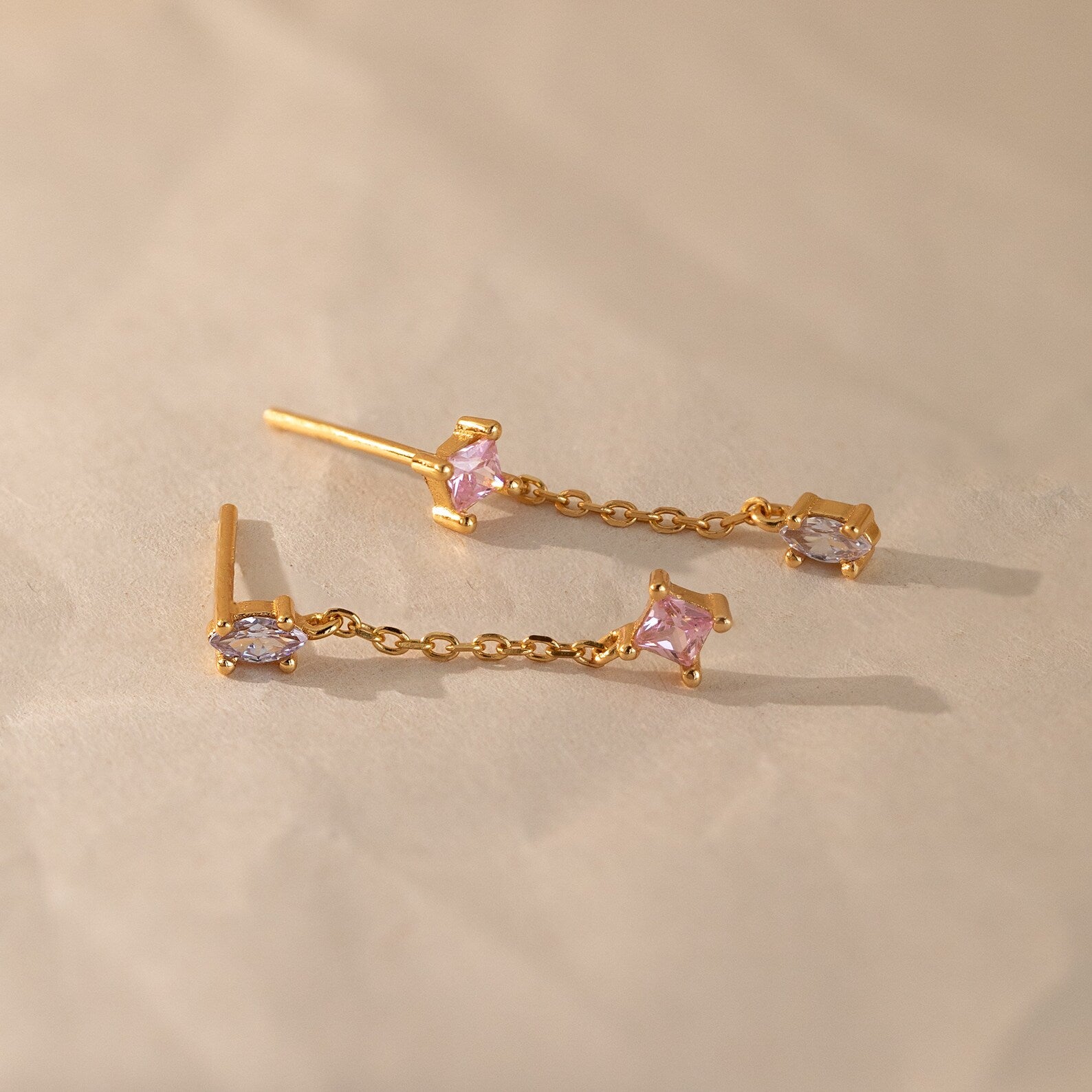 The Mixed Crystal Drop Earrings feature gold chain drops with pink and lavender gemstones, creating a minimalist style perfect for any occasion. These elegant dangling studs are shown on a light beige surface.