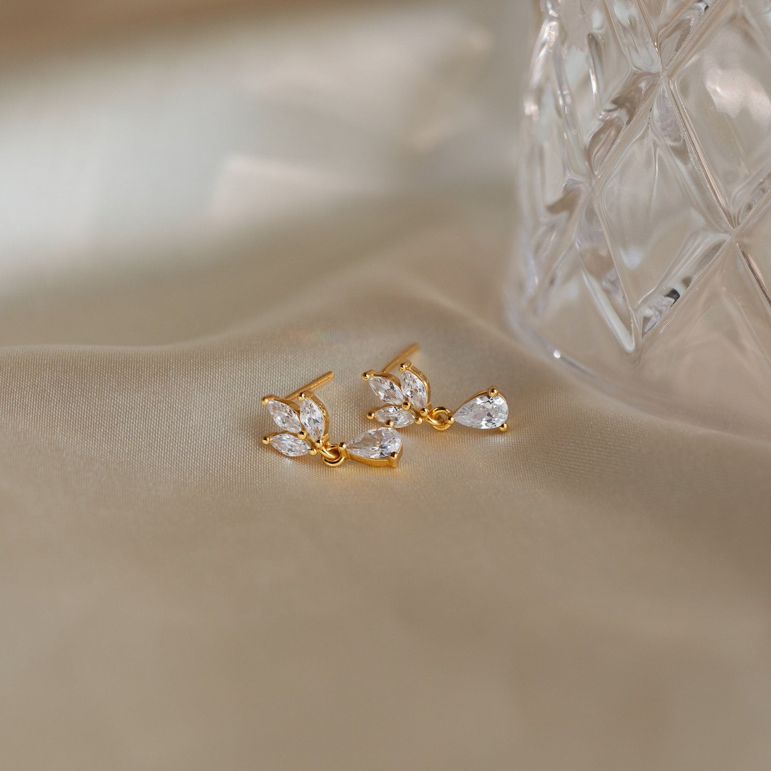 Marquise Diamond Drop Studs—gold earrings with clear, leaf-shaped gemstones inspired by Marquise-Cut Diamonds—rest on soft beige fabric beside a glass object.
