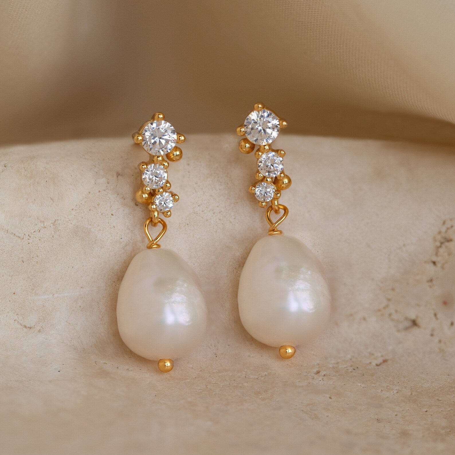 A close-up image of a pair of elegant drop earrings featuring three sparkling round-cut cubic zirconia stones set vertically in gold-toned prongs. Dangling below each cluster is a lustrous, organically shaped baroque freshwater pearl with a warm creamy tone