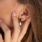 A hand touches an ear adorned with Emerald Pearl Drop Earrings, featuring a gold setting, green gem, and dangling pearl.