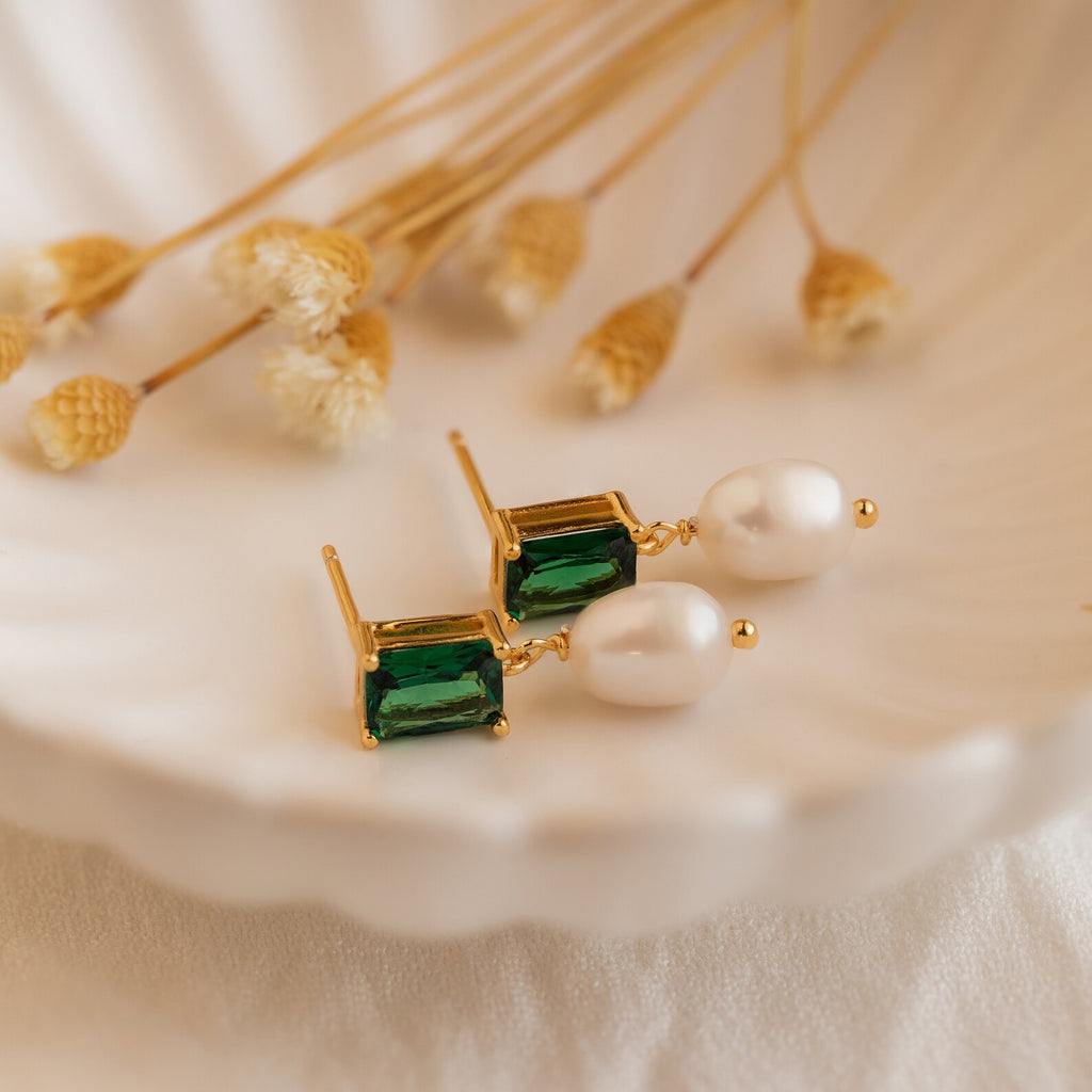 Emerald Pearl Drop Earrings with vintage green gemstones and white pearls are displayed on a scallop dish, accented by dried flowers in the background.