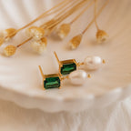 Emerald Pearl Drop Earrings with vintage green gemstones and white pearls are displayed on a scallop dish, accented by dried flowers in the background.