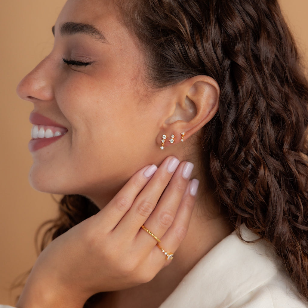 A woman with curly hair smiles, showcasing the Triple Diamond Studs Set and a gold ring on her hand.