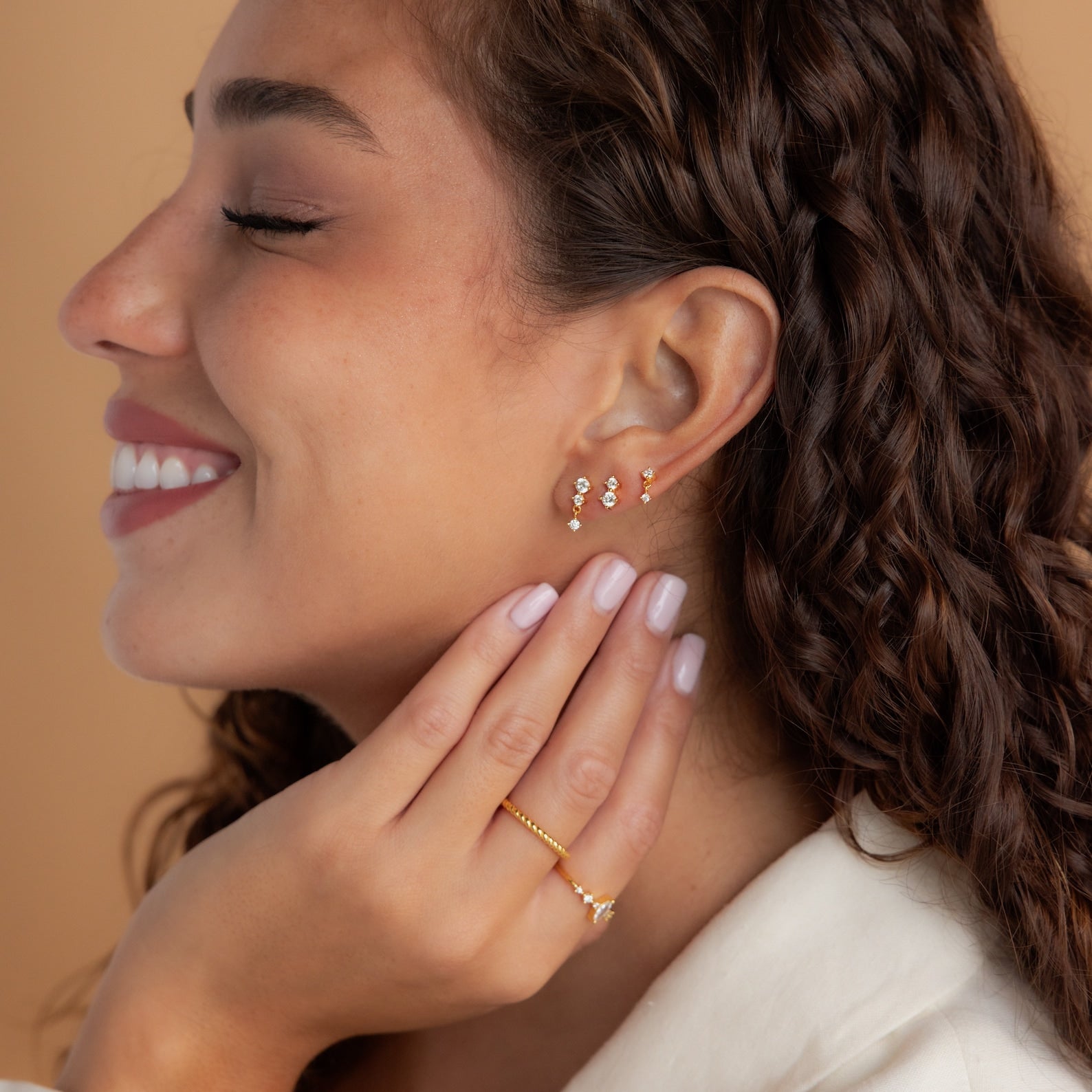 A woman with curly hair smiles, showcasing the Triple Diamond Studs Set and a gold ring on her hand.