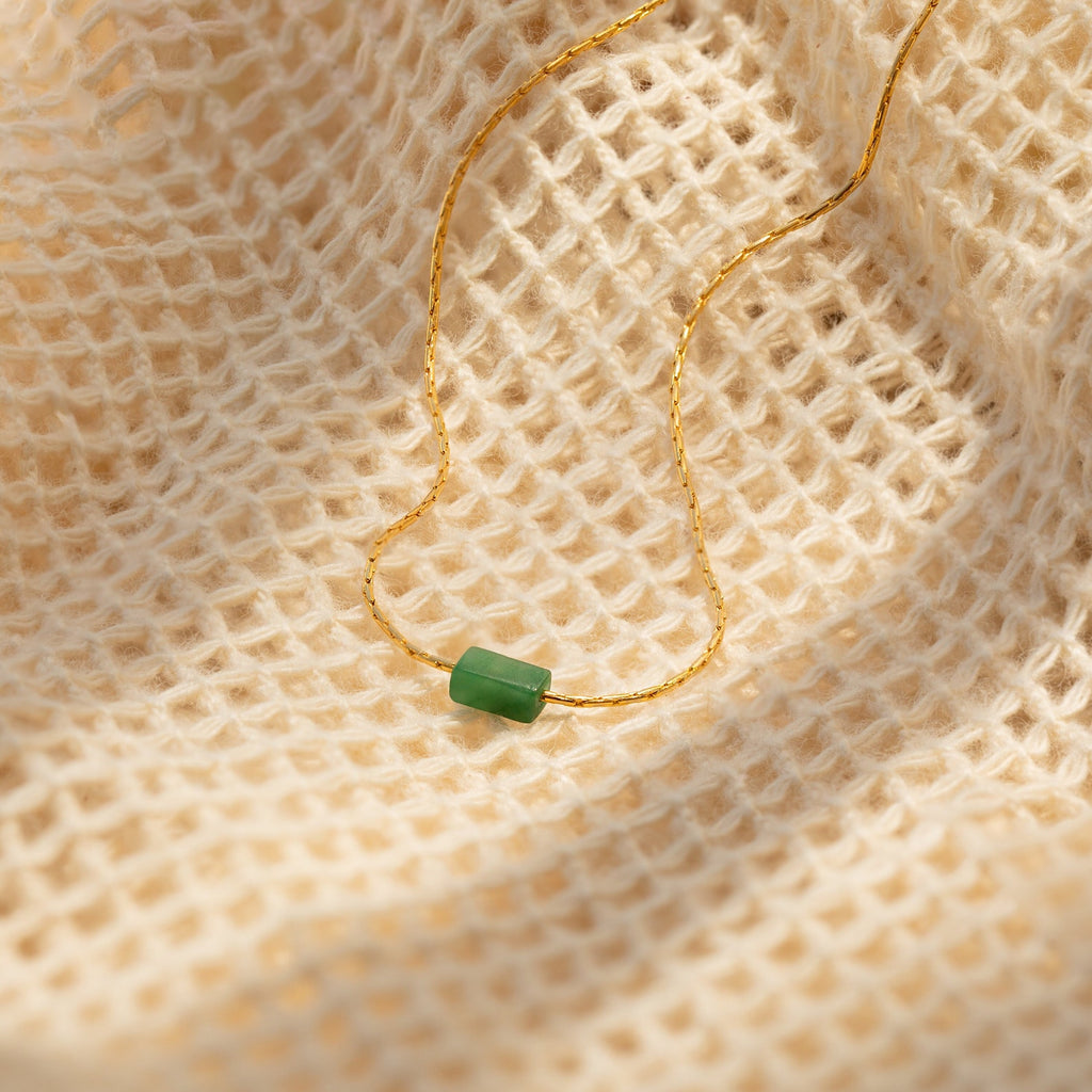 Minimalist gold necklace with a small horizontal jade bar pendant, displayed on a textured cream fabric background.