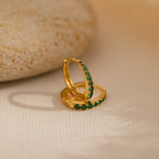 The Vera Emerald Birthstone Huggies feature elegant gold hoops with green gemstones, displayed on a light surface beside a smooth beige stone—perfect for adding a refined touch to any look.