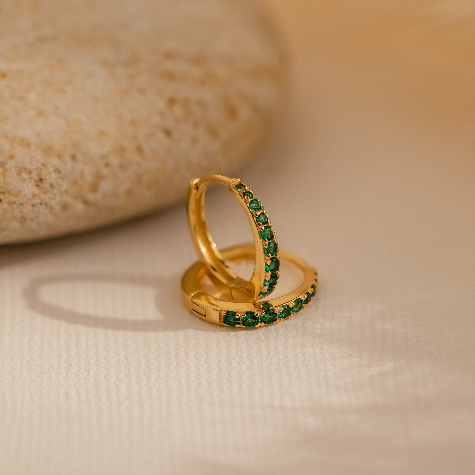 The Vera Emerald Birthstone Huggies feature elegant gold hoops with green gemstones, displayed on a light surface beside a smooth beige stone—perfect for adding a refined touch to any look.