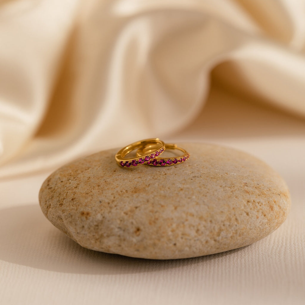 The Vera Ruby Birthstone Huggies, with their vibrant pink gemstones, rest on a smooth stone against a cream-colored fabric backdrop, reminiscent of colorful earrings.