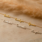 Three gold and three silver Dainty Tiny Multiple Name Necklaces with personalized nameplates are displayed on a light wooden surface beside brown pampas grass.