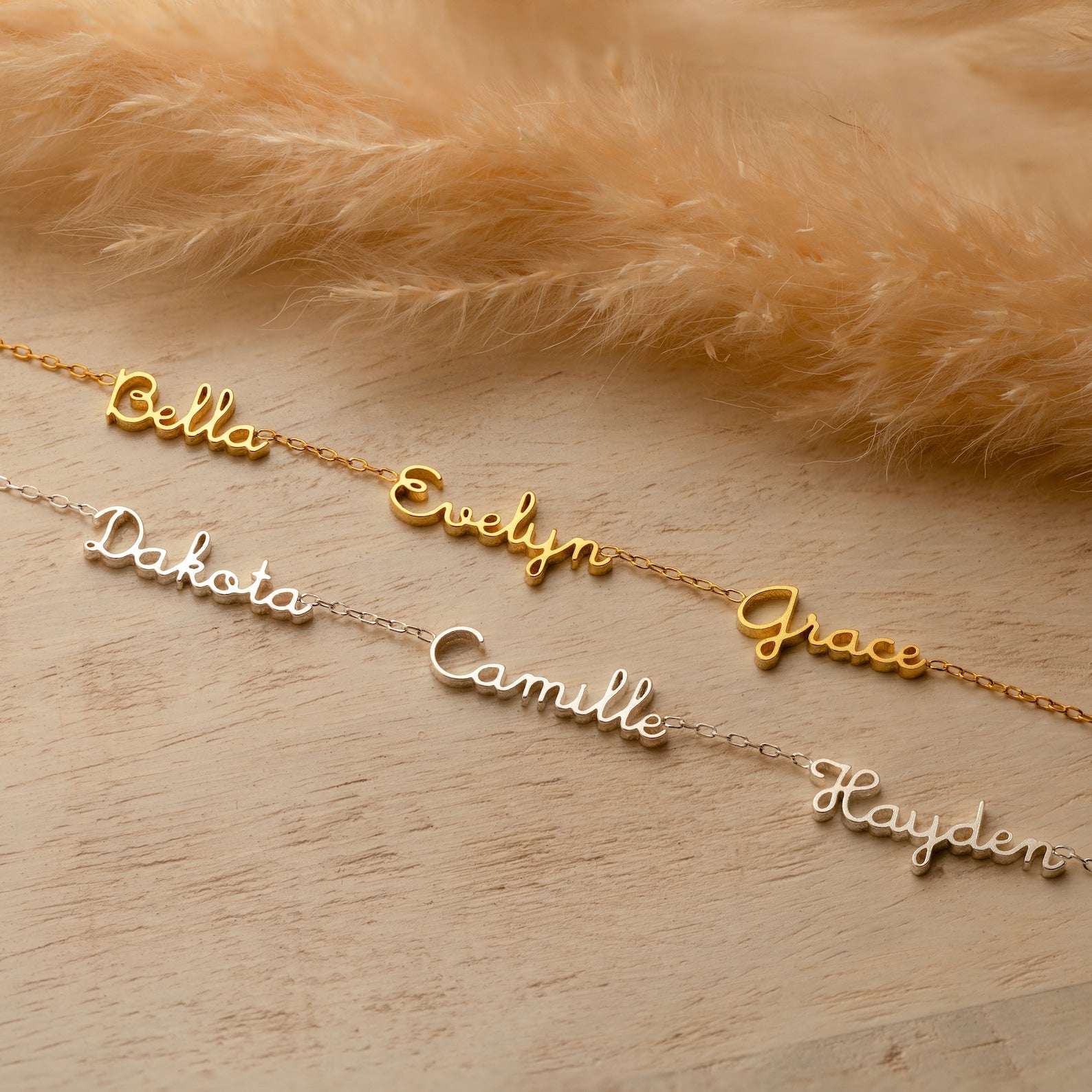 Three gold and three silver Dainty Tiny Multiple Name Necklaces with personalized nameplates are displayed on a light wooden surface beside brown pampas grass.