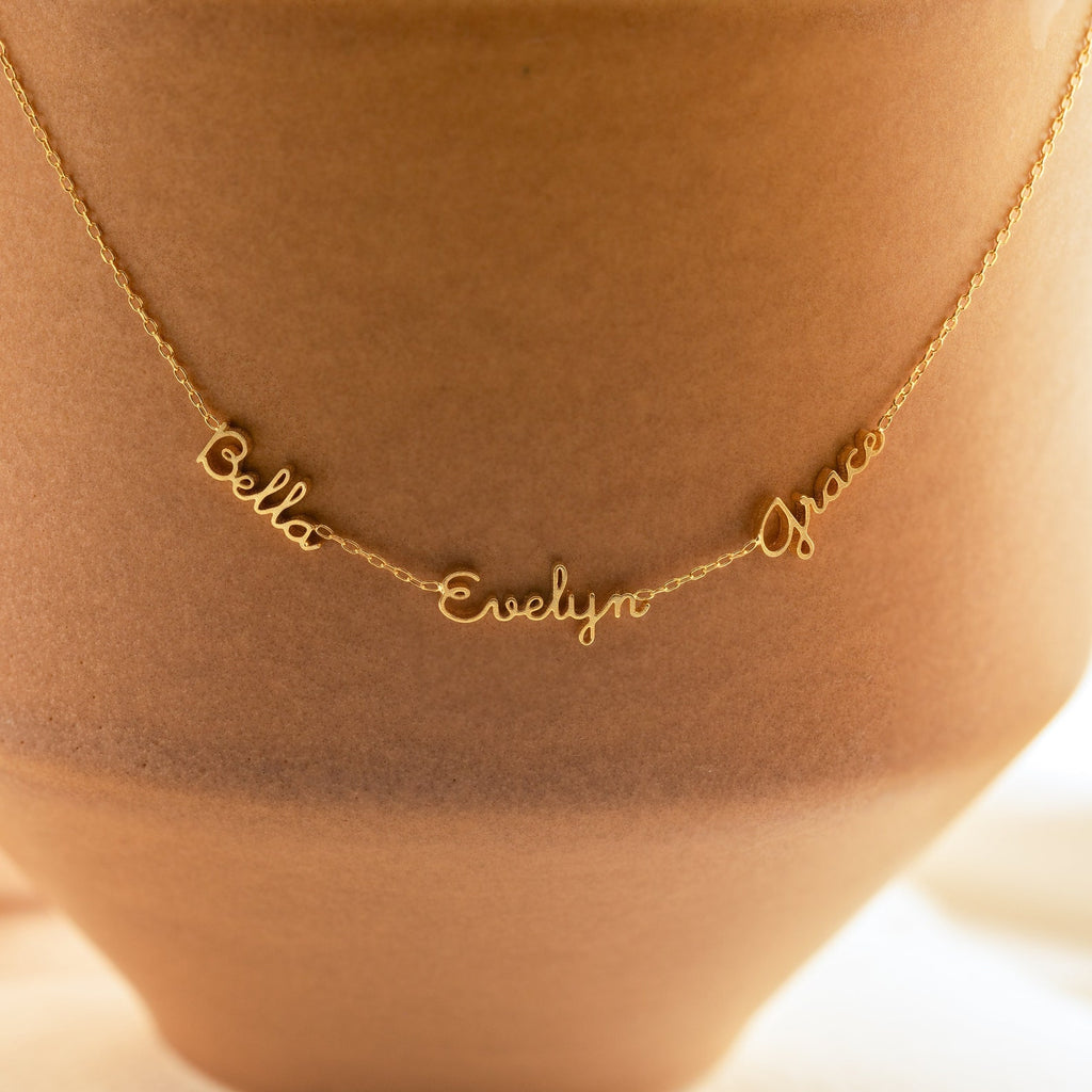 The Dainty Tiny Multiple Name Necklace features "Bella," "Evelyn," and "Grace" in elegant cursive, draped on a tan vase—making this personalized accessory a meaningful statement piece.