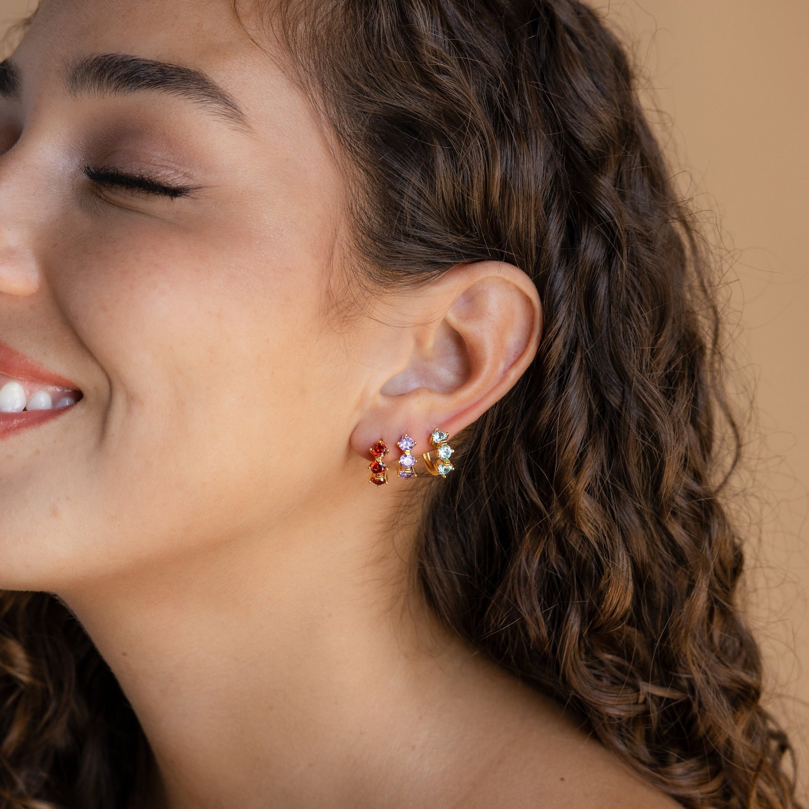 Woman with curly hair smiling, wearing vibrant Triple Opal Birthstone Huggies—perfect for those who love minimalist, customizable gemstone jewelry.