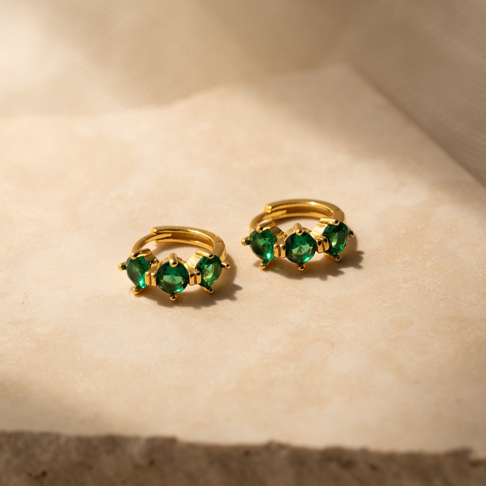 Triple Ruby Birthstone Huggies in gold, each adorned with three vibrant rubies, displayed elegantly on a beige stone surface—a perfect choice for lovers of personalized jewelry.