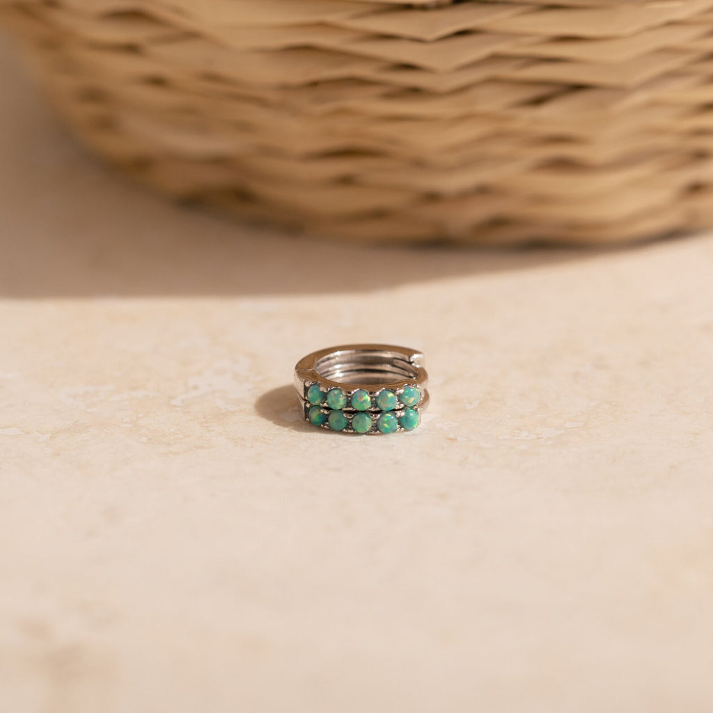 A silver ring with two rows of turquoise stones sits on a light surface near a woven basket—the perfect complement to your Garden Green Opal Huggies.
