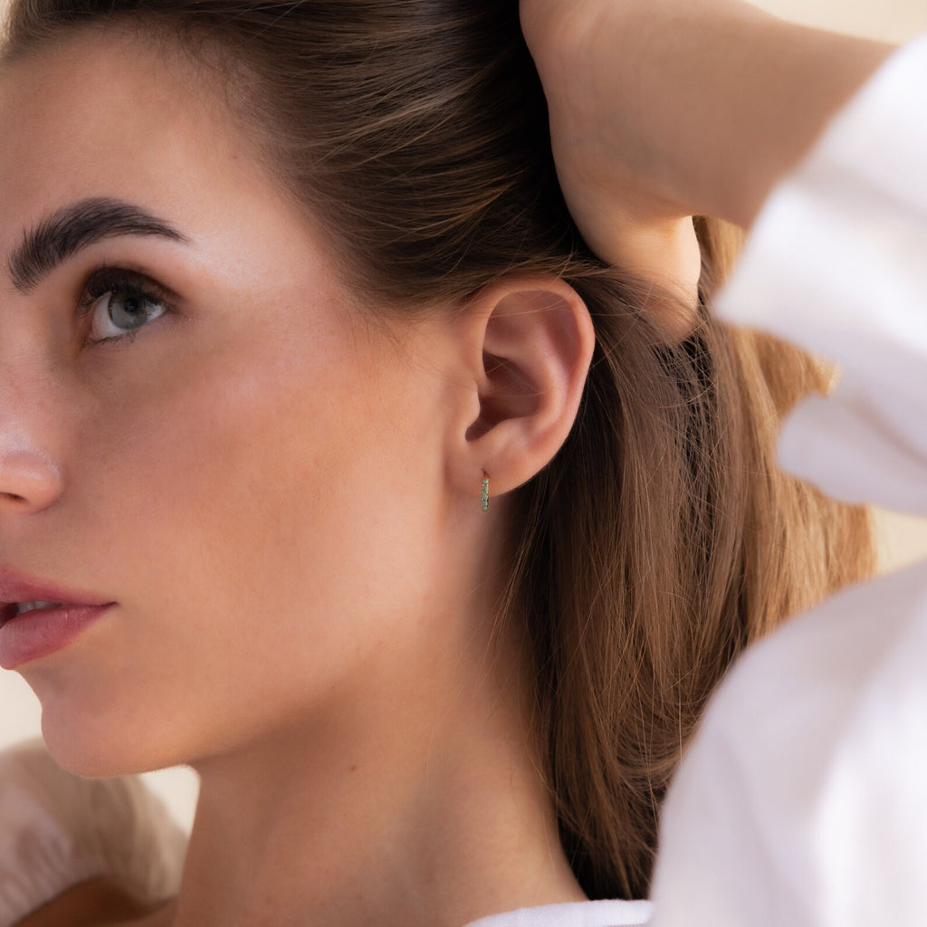 A woman with light skin and brown hair looks to the side, wearing Garden Green Opal Huggies—small green gemstone hoop earrings.