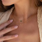 A woman wears two delicate gold necklaces, including the Dainty Aventurine Bar Necklace, featuring a green crystal bar pendant, as she touches her chest with a manicured hand.
