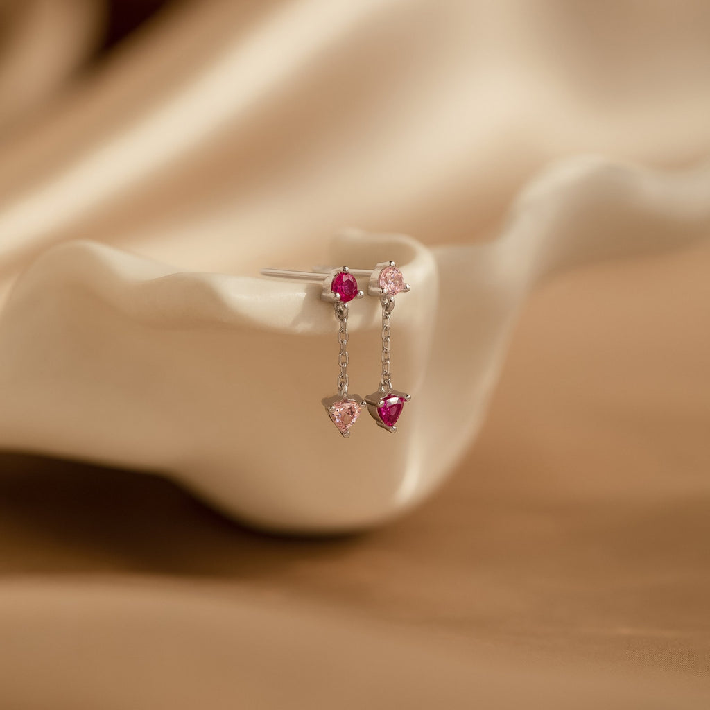 The Mixed Pink Drop Earrings, adorned with pink gemstones and displayed on a white shell, add a delicate and romantic touch to any outfit.