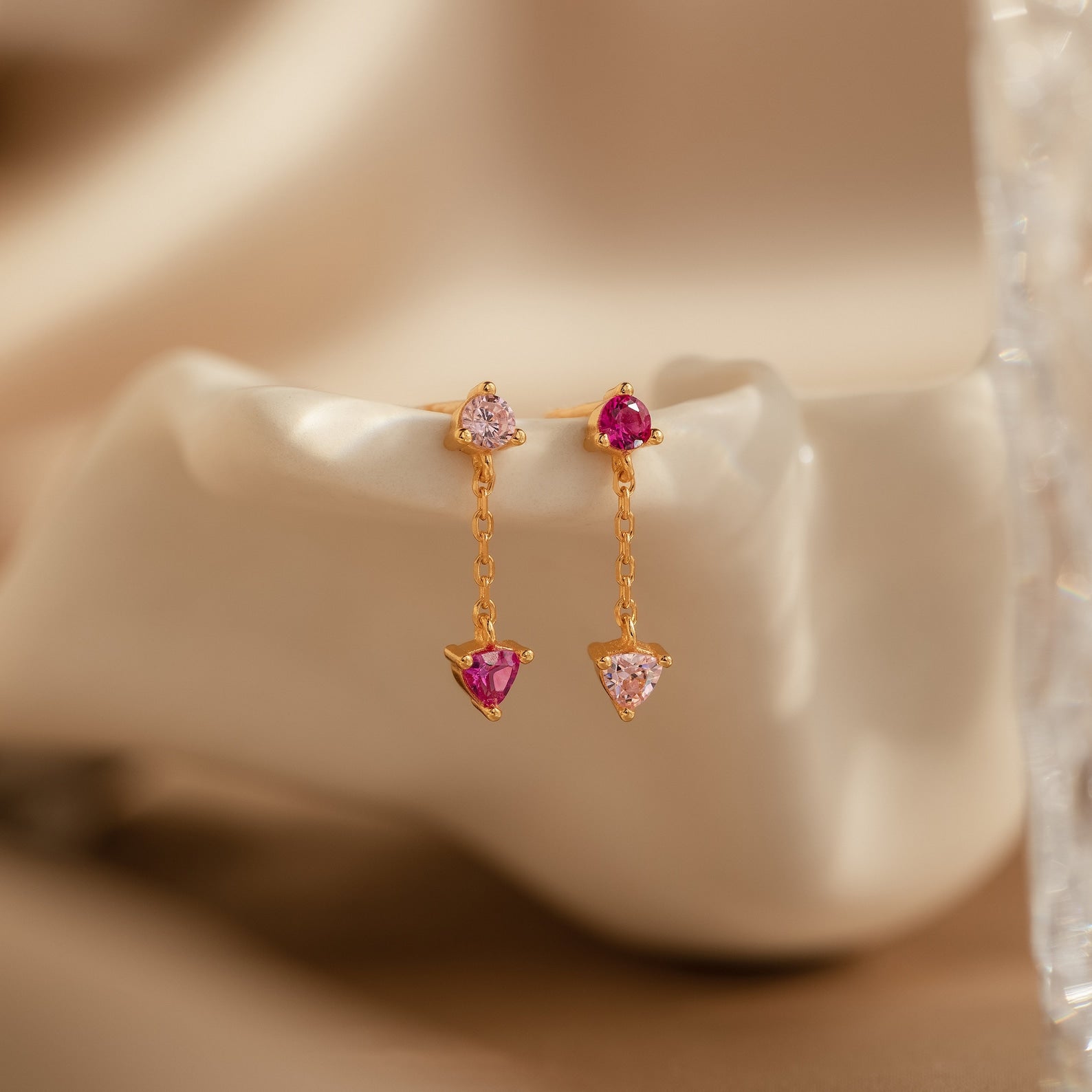 Mixed Pink Drop Earrings featuring gold-toned, mismatched pink drops with clear and pink gemstones in triangular settings and a delicate chain, displayed on a neutral background.