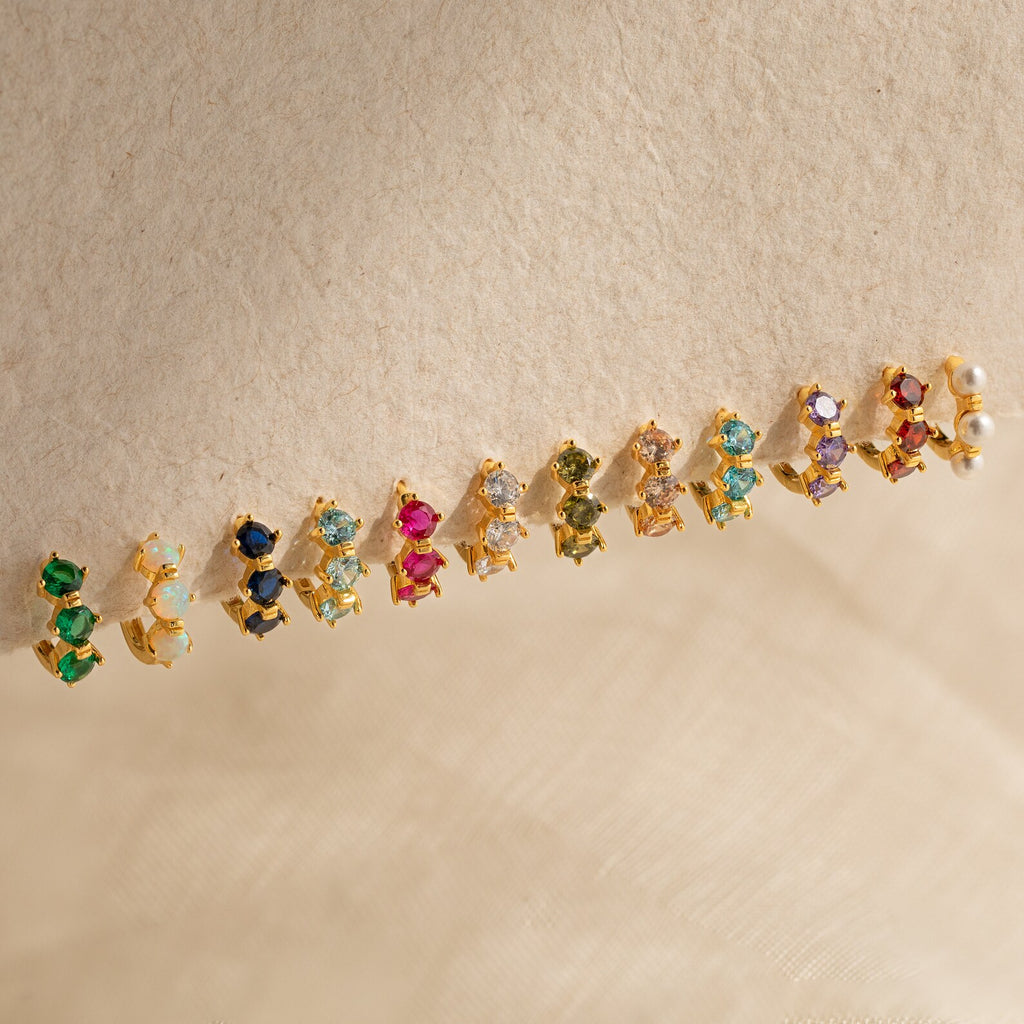 The Triple Ruby Birthstone Huggies, featuring three vibrant rubies set in gold, are perfect for fans of customizable or personalized jewelry and make a striking statement on a soft beige fabric background.